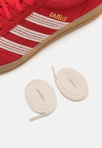 Röda mockaskor med tre vita textilränder, "Gazelle"-logotyp i gult och två vita bomullssnören bredvid.