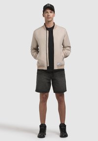 Jachetă bomber bej, tricou negru, pantaloni scurți din denim negri, adidași negri cu glezna înaltă și o șapcă neagră. Material neted, design minimalistic, fără modele.