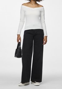 Vit off-shoulder långärmad topp i kombination med svarta vida jeans och svart handväska; modellen bär vita canvas sneakers.