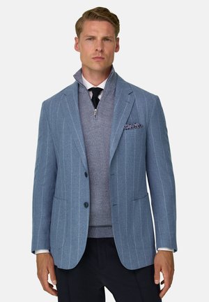 Homme blond portant un blazer bleu clair à fines rayures sur un pull gris à demi-fermeture éclair, une chemise blanche, une cravate noire et un pantalon foncé.