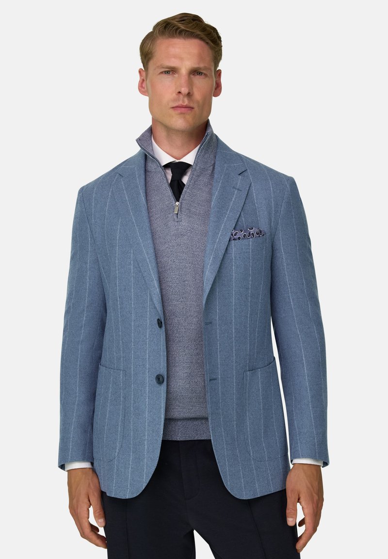 Homme blond portant un blazer bleu clair à fines rayures sur un pull gris à demi-fermeture éclair, une chemise blanche, une cravate noire et un pantalon foncé.