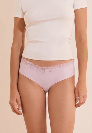Mujer con ropa interior rosa claro con encaje y una camiseta corta blanca, de pie frente a un fondo beige liso.