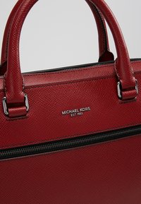 Sac à main en cuir rouge texturé avec double poignée, fermeture éclair argentée et logo métallique "Michael Kors EST 1981" à l'avant.