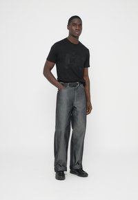 Μαύρο γραφικό Τ-shirt με διακριτικό λογότυπο, συνδυασμένο με γκρι τζιν παντελόνι φαρδιάς γραμμής και μαύρα παπούτσια, με ομαλή υφή και καθαρές γραμμές.