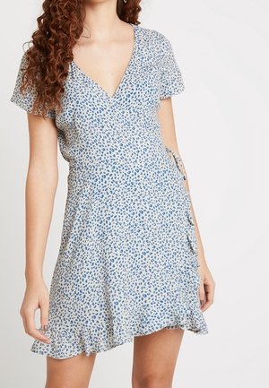 Kobieta w krótkim rękawie, sukience kopertowej z dekoltem w serek, w niebiesko-biały wzór w panterkę i z falbaną u dołu.