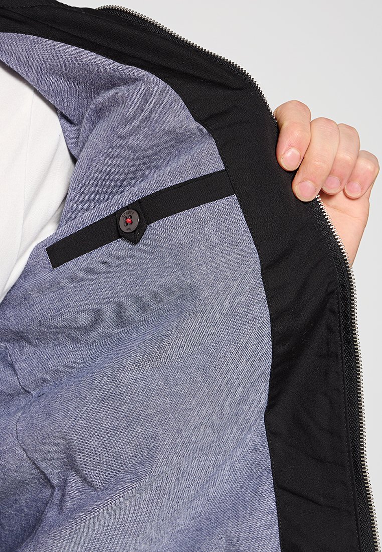 Intérieur d'une veste noire avec une doublure en tissu gris. Les caractéristiques comprennent une sangle noire avec un bouton et une fermeture éclair argentée le long du bord.