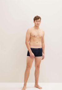 Schwarze Baumwollboxershorts mit einem weichen Bund, der "TOM TAILOR" in kontrastierender Farbe zeigt. Model steht vor einem neutralen Hintergrund.