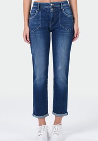 Femme portant un jean en denim bleu retroussé avec des poches avant et des baskets blanches, se tenant devant un fond neutre.