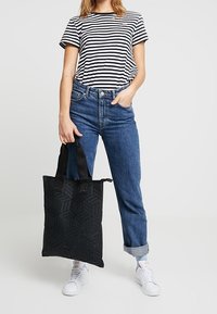 Sac cabas noir avec un motif géométrique, doté de poignées robustes, tenu devant le modèle portant un t-shirt rayé et un jean bleu.