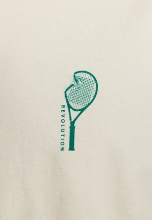 T-shirt en coton beige avec un graphisme vert d'une raquette de tennis cassée accompagnée du mot "RÉVOLUTION" imprimé verticalement.