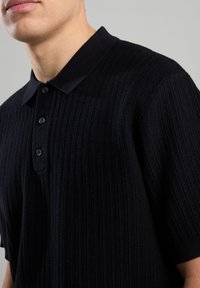 Schwarzes geripptes Poloshirt mit klassischem Kragen, Knopfleiste mit drei Knöpfen und kurzen Ärmeln. Hergestellt aus strukturierter Strickware.