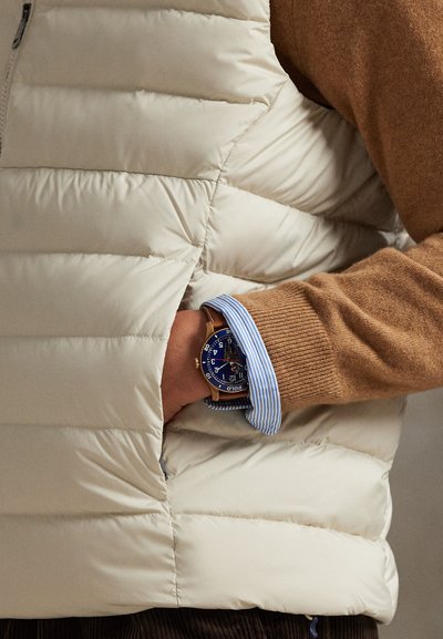 Lätt cremefärgad vadderad väst med quiltdesign, bärs över en brun tröja. Blå randig skjorta som skymtar under. Guldur med blå urtavla.