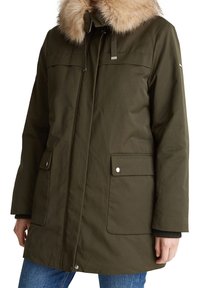 Parka d'hiver vert olive avec capuche bordée de fourrure, poches avant à pression, portée sur un jean bleu sur une personne.