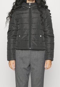 Svart puffjacka med dragkedja framtill, horisontella sömmar och hög krage. Har sidofickor och en slät, glänsande textur.