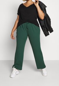 Svart draperad topp med korta ärmar, matchad med gröna vida byxor med dragsko. Modellen bär vita sneakers och håller en svart jacka.