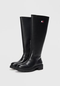 Bottes noires en cuir hauteur genou avec une fermeture éclair et une semelle extérieure texturée. Présente un patch logo en haut avec des accents rouge, blanc et bleu.