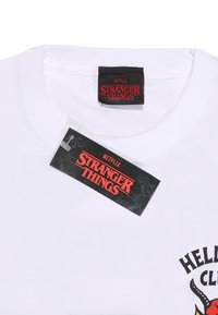 Bílý bavlněný tričko s černou a červenou nášivkou "Stranger Things", s dekorativními grafikami a připojenou visačkou.