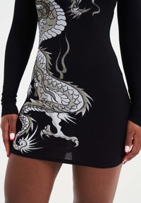 Schwarzes geripptes Kleid mit metallischen Drachen-Graphics in Silber und Gold. Lange Ärmel und eine taillierte Silhouette. Glatte Textur mit lebhaften Details.