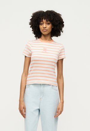 Frau mit lockigem Haar, die ein weißes und pfirsichfarben gestreiftes T-Shirt mit kurzen Ärmeln und hellblaue, hoch taillierte Jeans trägt, steht vor einem einfarbigen Hintergrund.