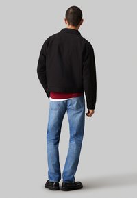 Giacca nera slim fit, abbinata a jeans blu. La giacca presenta maniche lunghe e un taglio dritto, evidenziando un design minimalista.
