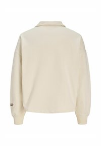 Μακρυμάνικο πουκάμισο σε κρεμ χρώμα με γιακά, ριμπ μανσέτες και ορατή μικρή λεπτομέρεια κειμένου στο αριστερό μανίκι κοντά στον καρπό.