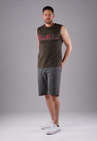 Sleeveless olive tank top with red graphic, paired with gray denim shorts. Model wearing white sneakers stands in a neutral background.