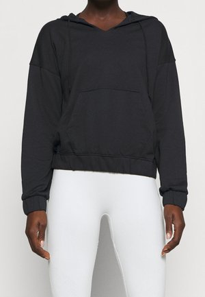 Personne portant un sweat à capuche noir court avec une poche avant et des leggings blancs moulants sur un fond uni.