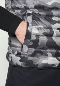 Main dans la poche d'un gilet matelassé camouflage gris et noir porté par-dessus une chemise noire à manches longues et un pantalon noir.