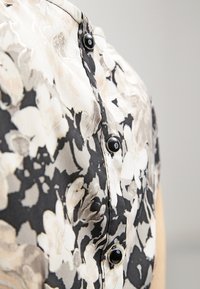 Gros plan d’un chemisier floral noir et blanc avec des boutons noirs à l’avant sur un fond uni.