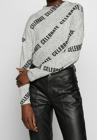 Grå långärmad tröja med svart tryckt text "CELEBRATE" diagonalt; i kombination med svarta läderbyxor som visar en jämn textur och passform.