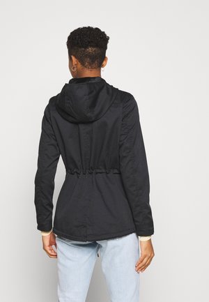 Personne aux cheveux courts et bouclés portant une veste à capuche noire et un jean bleu clair, debout de dos sur un fond uni.