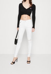 Cárdigan corto de canalé negro con cierre de botón, combinado con jeans ajustados blancos con textura de canalé y rotura en la rodilla. Tacones abiertos negros.