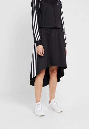 Persoon draagt een zwart Adidas trainingsjack met witte strepen en een bijpassende high-low rok, gecombineerd met witte sneakers, tegen een witte achtergrond.