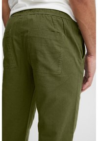 Mies, jolla on päässään oliivinvihreät housut, joissa on joustava vyötärönauha ja taskut, kuvattuna takaa vyötäröstä reiden keskikohdalle.