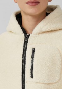 Helle Fleece-Hoodie in Cremefarbe mit einem schwarzen Reißverschluss, einer Fronttasche und einer strukturierten Oberfläche. Die Kapuze und der Kragen sind sanft geformt.