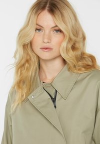 Hellgrüner Trenchcoat mit einem Kragen, ausgestattet mit einem Reißverschluss und Druckknöpfen. Glattes Gewebe mit einem schlichten Design und ohne sichtbare Muster.