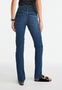 Tumed pesu denimist valmistatud laienevad teksad keskmise kõrgusega vöökoht, millel on tavaline tagatasku disain ja sile tekstuur. Kandmise ajal mustade sandaalidega.