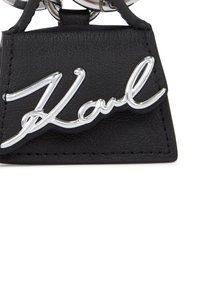 Zwarte leren sleutelhanger met zilveren "Karl" script logo, voorzien van een textuur en een driehoekige vorm.
