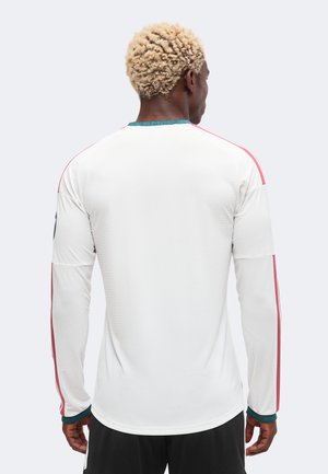 Person med krøllet blondt hår iført en hvid langærmet sportstrøje med røde striber på ærmerne og sorte shorts, set bagfra.