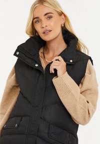 Gilet a vento nero con colletto alto e chiusura a zip, caratterizzato da ampie tasche frontali e una texture trapuntata. Indossato sopra un maglione a maglia beige.