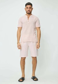 Chemise en lin rose clair à manches courtes avec col cranté et boutons, associée à un short assorti rose clair et des sandales plates noires.