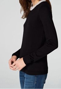 Svart långärmad tröja med en texturerad pärlkrage. Har en figurnära design och ribbad nederkant, i kombination med blå jeans.