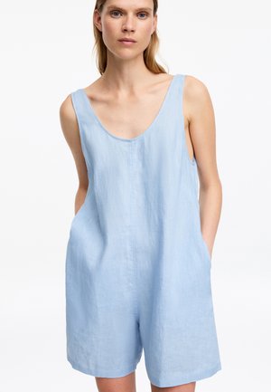 Femme portant un combishort en lin bleu clair sans manches avec un profond décolleté en U et les mains dans les poches, fond blanc.