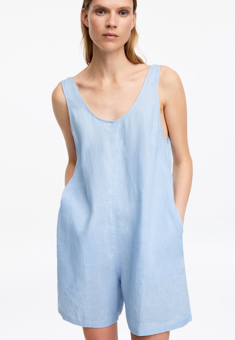 Femme portant un combishort en lin bleu clair sans manches avec un profond décolleté en U et les mains dans les poches, fond blanc.