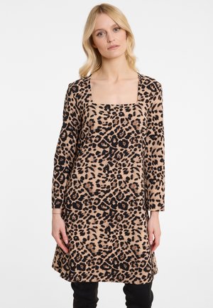 Frau in einem knielangen, langärmligen Kleid mit Leopardenmuster und quadratischem Ausschnitt, stehend vor einem schlichten weißen Hintergrund.