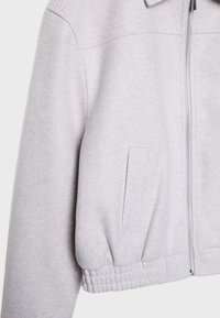 Veste gris clair en tissu doux et texturé ; dotée d'une fermeture éclair, d'un col et d'une taille élastiquée avec un détail de poche latérale.