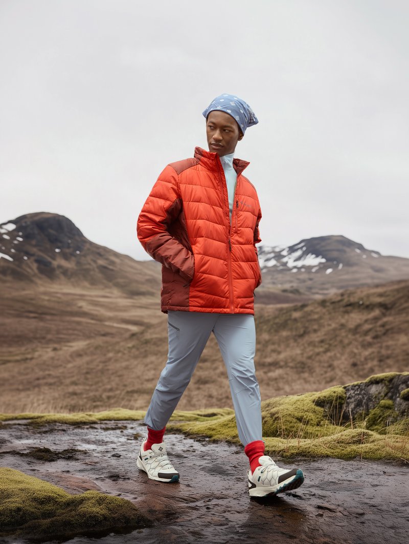 Czerwona kurtka puchowa z wysokim kołnierzem, jasnoniebieska koszula z długim rękawem, jasnoszare spodnie, czerwone skarpetki i białe sneakersy z akcentami w kolorze teal.