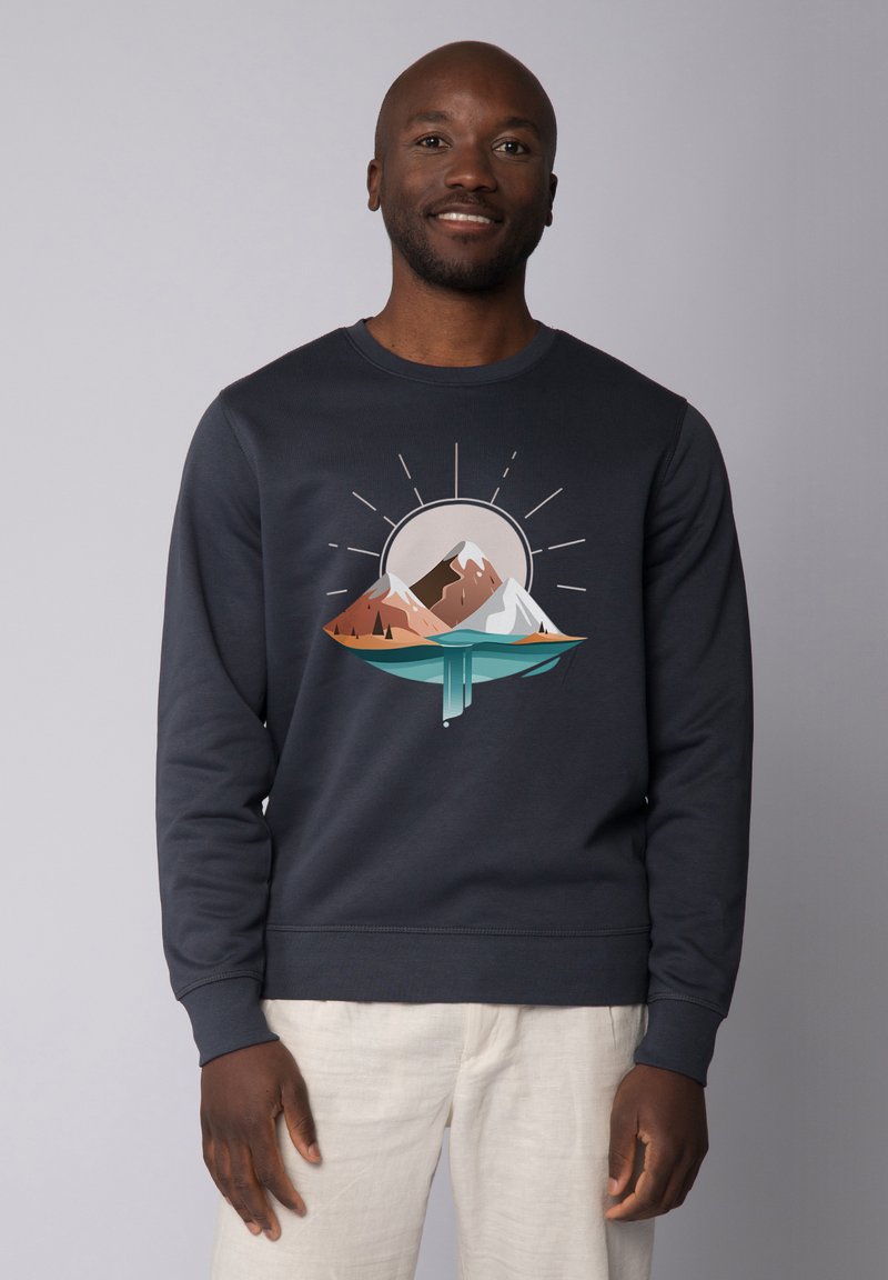 Marineblauwe sweatshirt met een bergafbeelding met bruine en witte toppen boven een teal kleurig water, versterkt door zonnestralen. Zachte textuur.