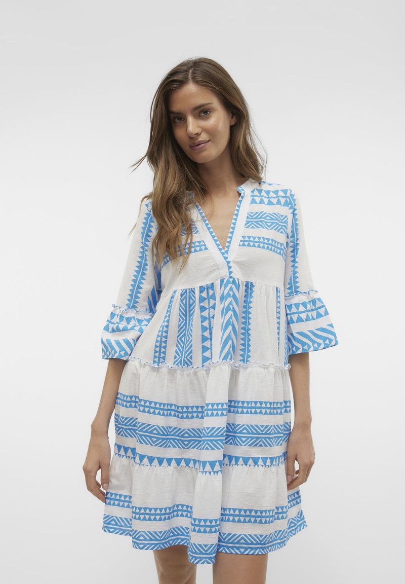 Robe blanche avec des motifs géométriques bleus variés, dotée d'un décolleté en V, d'un design en plusieurs niveaux et de manches évasées. Tissu doux et texturé.