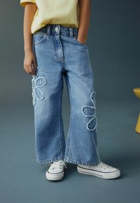 Šviesiai mėlynos plačios kojinės džinsai su paberztais gėlių siuvinėjimais ant kelių, derinami su baltais sportbačiais ir geltona trumpomis rankovėmis palaidine.